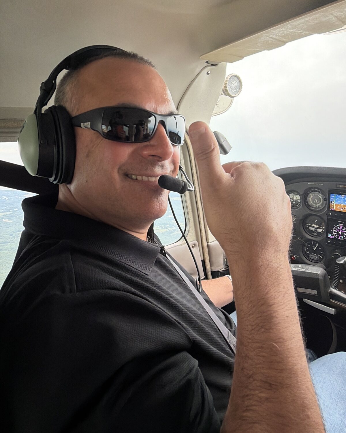 Greg Boghosian in a cockpit wearing a headset and sunglasses, giving a thumbs up